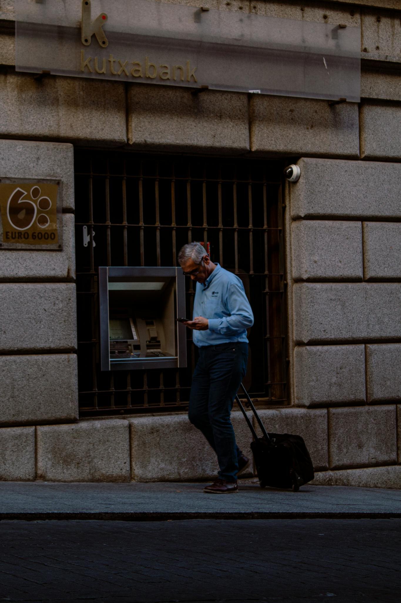 A businessman using an ATM on city streets while checking his phone.