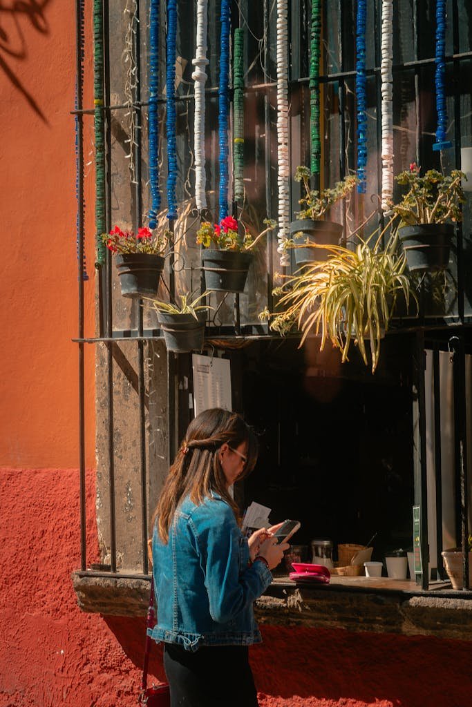 A woman stands by a vividly decorated window in San Miguel de Allende, Mexico, bathed in sunlight.